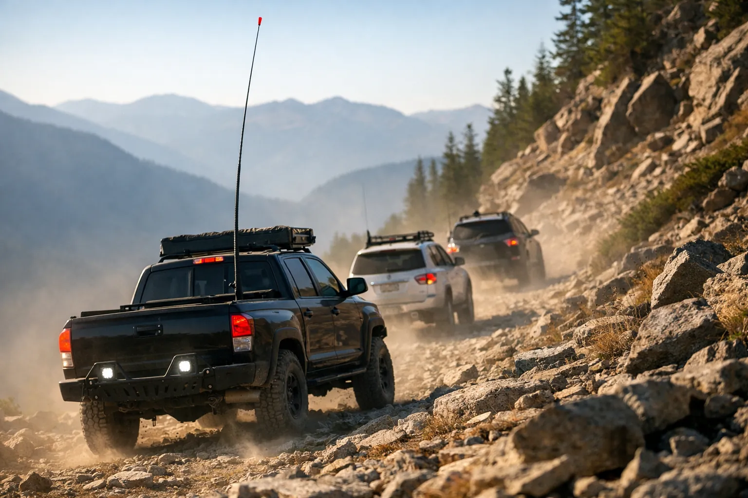 A line of off-road vehicles on a rocky trail, the lead vehicle with a tall whip antenna and others with short antennas
