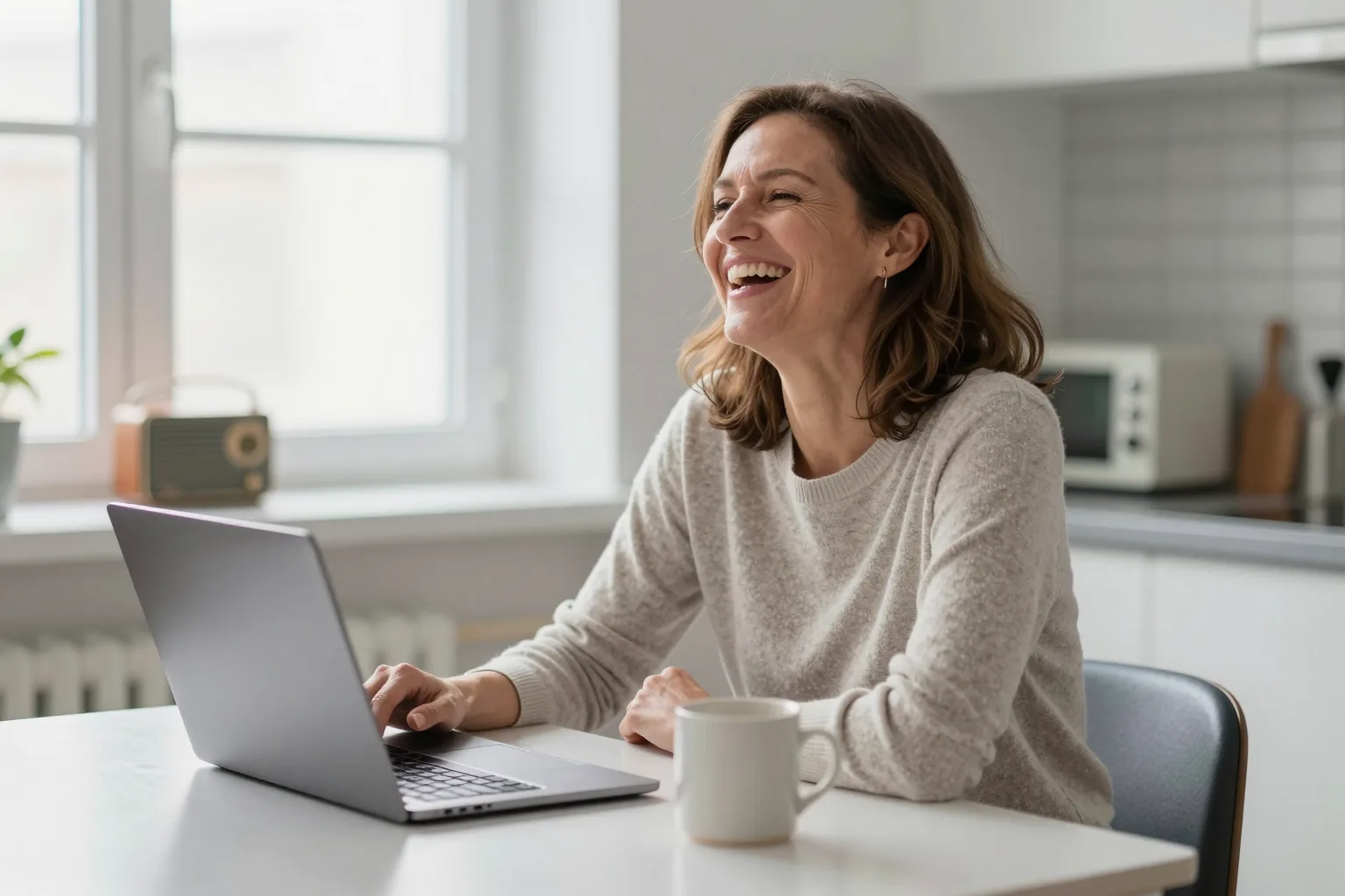 A woman at a bright kitchen table laughing as she works on her laptop, a coffee mug beside her and a small radio on the windowsill